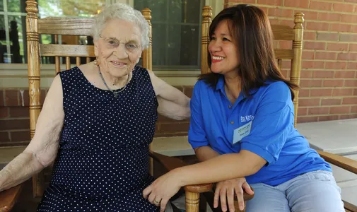 Smiling resident with smiling caregiver on the front porch in rocking chairs.